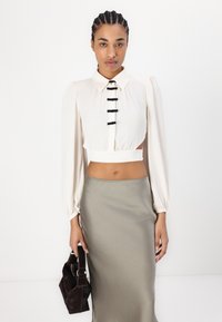 Eine cremefarbene Bluse mit Puffärmeln, schwarzen Knopfdetails und cropped Saum, kombiniert mit einem silbernen Rock, gehalten mit einer strukturierten dunklen Handtasche.