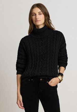 Junge Frau trägt einen schwarzen Zopfpullover mit Rollkragen, schwarze Hose, eine schwarze Uhr und ein goldenes Manschettenarmband, steht mit einer Hand in der Tasche.