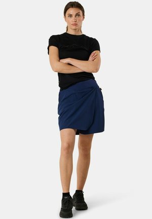 Femme debout, les bras croisés, portant un t-shirt noir à manches courtes, une jupe portefeuille bleu marine, des chaussettes noires et des chaussures de sport noires.