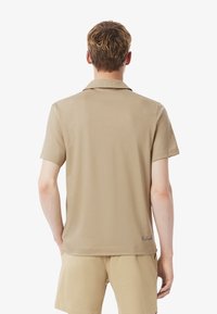 Beige kortärmad pikétröja med en nedfälld krage och en slät textur, med en subtil logodetalj vid fållen.