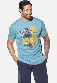 Smiling man with beard, wearing a light blue "Northern Horizons" graphic t-shirt and dark blue shorts, hands in pockets, showing arm tattoos.