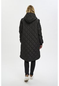 Schwarzer langer gepolsterter Mantel mit Rautensteppung, Kapuze und Reißverschlusstaschen, kombiniert mit schwarzen Leggings und weißen Sneakers.