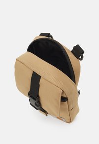 Saco de tela bege com um zipper preto, alças ajustáveis e uma fivela de liberação rápida. O interior apresenta um forro preto. Design compacto.