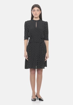 Donna che indossa un vestito nero fino al ginocchio con pois bianchi, maniche a metà braccio, scollo a goccia e scarpe nere con tacco basso, ferma.