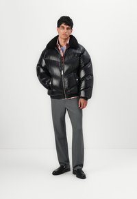 Zwarte pufferjack met een glanzende afwerking, ritsluiting en een zwarte bontkraag, gecombineerd met grijze broek en zwarte schoenen.