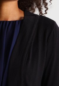 Primer plano de una persona que lleva un vestido azul marino con escote fruncido y un cárdigan negro texturizado, con el cabello rizado visible.