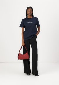 Marine T-skjorte med "Calvin Klein" logo, kombinert med svarte vide bukser og en rød lærveske. Enkelt, moderne design med komplementære farger.