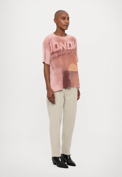 T-shirt graphique rose avec le texte "BONDY" et un motif de coucher de soleil ; coupe décontractée, manches courtes. Assorti avec un pantalon clair et des bottines noires.
