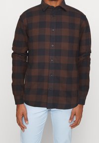 Bruine en zwarte geruite blouse met lange mouwen, knoopsluiting aan de voorkant en subtiele textuur. Voorzien van donkere knopen en een klassieke kraag.