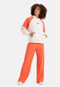 Hooded sweatshirt in cream with orange raglan sleeves, featuring a small logo and striped accents. Paired with orange ribbed wide-leg pants.