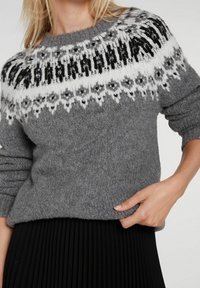 Pull gris en tricot avec empiècement noir et blanc à motifs, poignets côtelés et coupe ample. Associé à une jupe noire plissée.