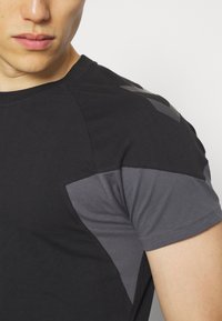 Gros plan d'une personne portant un t-shirt de sport noir et gris à manches courtes avec un logo subtil sur l'épaule.