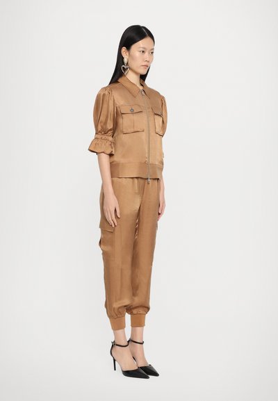 Bruine satijnen outfit met een ritssluiting jack met pofmouwen en twee zakken, gecombineerd met taps toelopende broek en zwarte high heels.