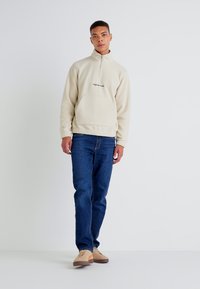 Light beige fleece pullover with a half-zip, front pocket, and black text logo. Paired with dark blue denim jeans and tan sneakers.