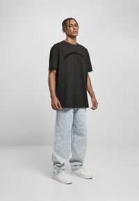 T-shirt en coton noir avec "URBAN CLASSIC" en texte courbé, associé à un jean baggy bleu clair et des baskets blanches. Modèle posant sur un fond neutre.