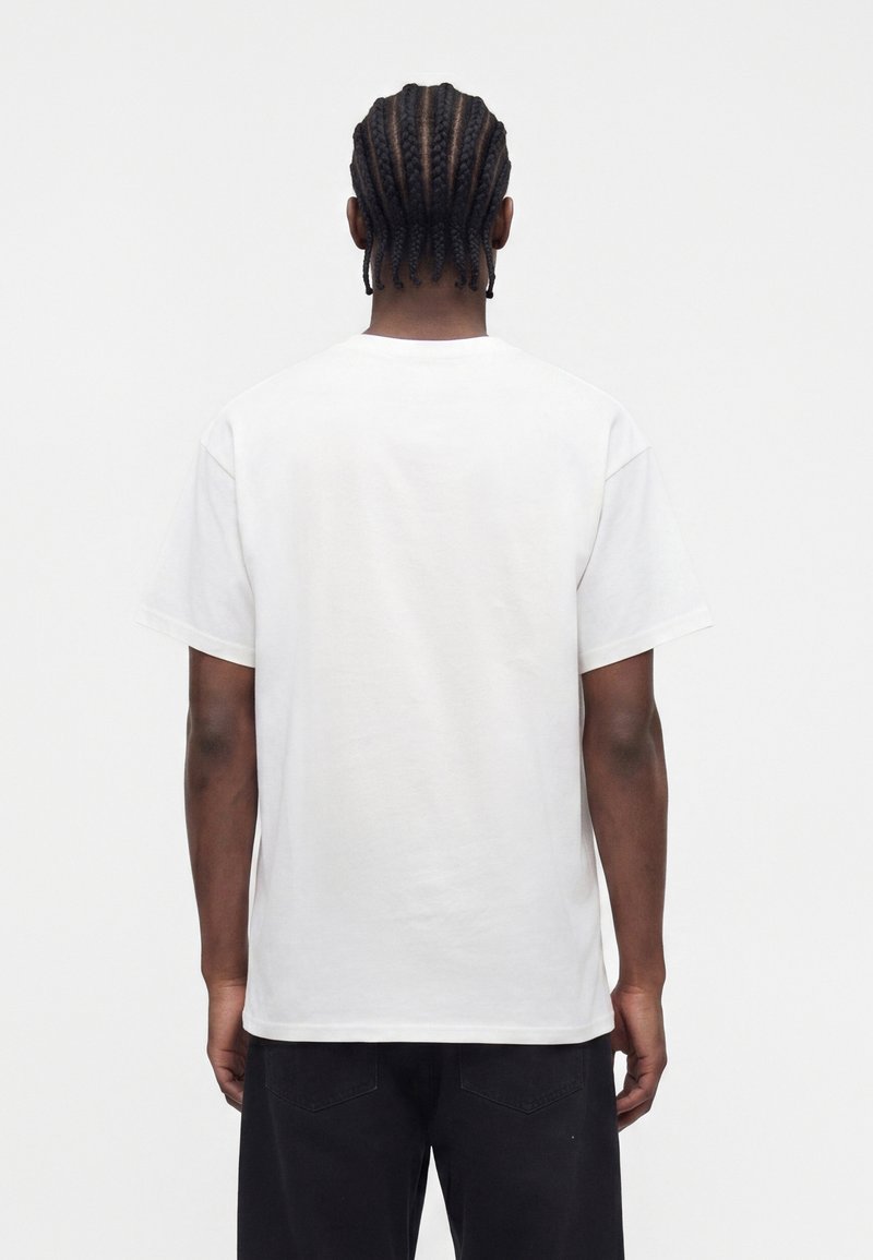 Mann mit Cornrow-Zöpfen, der ein schlichtes weißes T-Shirt mit kurzen Ärmeln und schwarze Hose trägt, von hinten vor weißem Hintergrund dargestellt.