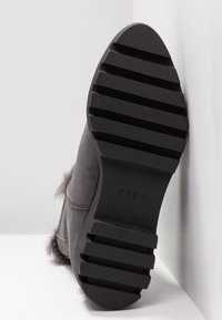 Semelle d'une botte grise avec des rainures horizontales profondes et un marquage "DKNY", inclinée contre un mur d'angle blanc.