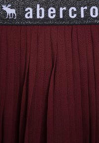 Czarny elastyczny pas w talii z białym logo w postaci łosia oraz napisem 'abercrombie' ponad materiałem marszczonej spódnicy w kolorze ciemnoczerwonym.