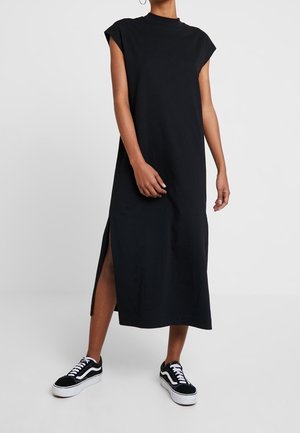 Kobieta w czarnej, bezrękawnikowej sukience midi z bocznym rozcięciem oraz czarnych niskich trampkach z białą podeszwą i sznurówkami.