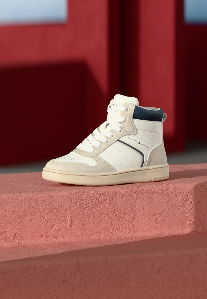 Valged kõrged tenniseketsid, mis on valmistatud sametist ja nahast, purpurse aktsent pahkluul ning tekstuurne tallaosa. Märkimisväärne brändi logo külgedel.