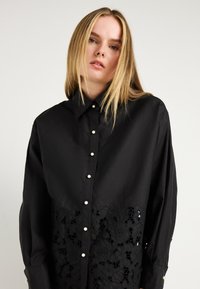 Camisa preta de botão com um design de renda floral na parte inferior, com botões semelhantes a pérolas e mangas longas e largas. Textura suave.