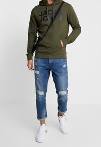 Grön hoodie med en stor svart logotyp, tillsammans med slitna blå jeans och vita sneakers. Outfiten inkluderar en svart axelväska.