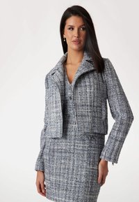 Harmaa ja sininen tweed-takki, jossa on lyhyt leikkaus, terävä kaulus ja vetoketjukiinnitys. Takissa on teksturoitu kangas ja muotoillut hihat.