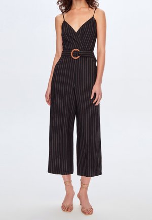 Kobieta ubrana w czarny kombinezon w prążki bez rękawów z szerokimi nogawkami, dekoltem w serek i paskiem z okrągłą klamrą, zestawiony z cielistymi sandałami na obcasie.