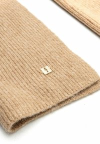 Beżowy, żeberkowy szalik dzianinowy wykonany z miękkiego materiału, z małym złotym akcentem. Krawędzie starannie wykończone, co zapewnia schludny wygląd.