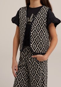 Czarna i biała kamizelka w wzory z rombami, z teksturowanej tkaniny, z zaokrąglonym kołnierzem, noszona na czarnej koszulce z marszczonymi rękawami.