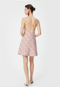 Witte jurk met een bloemenpatroon met rode bloemen en groene bladeren. Spaghetti bandjes met een kruisvormige achterkant. Korte A-lijn snit.