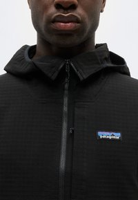 Κοντινό πλάνο ενός ατόμου που φοράει μαύρο μπουφάν Patagonia με φερμουάρ στο μπροστινό μέρος και κουκούλα, με το λογότυπο στο αριστερό μέρος του στήθους.