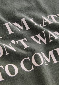 Un tissu vert froissé arbore un texte blanc audacieux disant "JE SUIS EN RETARD, JE NE VEUX PAS VENIR." L'accent est mis sur le texte et la texture.