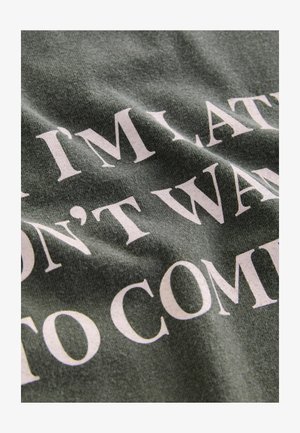 Un tessuto verde sgualcito presenta un bold testo bianco che dice "SONO IN RITARDO, NON VOGLIO VENIRE." L'attenzione è focalizzata sul testo e sulla texture.