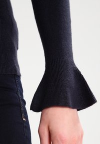 Pull bleu marine à manches évasées, confectionné en tissu tricoté doux. Présente une texture côtelée au niveau du poignet. Gros plan sur la manche et la main.