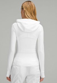 Un sweat à capuche blanc ajusté avec une texture douce, des manches longues et une capuche arrondie, doté d'un détail de couture dans le dos et d'ouvertures pour les pouces dans les poignets.
