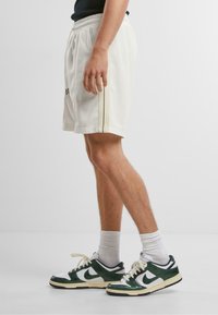 Pantaloni sportivi bianchi con vita elastica, caratterizzati da due strisce nere verticali lungo i lati. Abbinati a sneaker verdi e bianche.