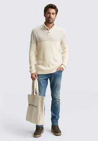 Kremowy prążkowany sweter z kołnierzykiem na guziki, w połączeniu z jasnymi dżinsami i brązowymi botkami, trzyma beżową torbę typu tote z logo.