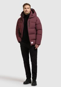 Bordeaux pufferjakke med hætte, lynlås foran og sidelommer. Båret over en sort turtleneck og sorte bukser, med sorte sko.