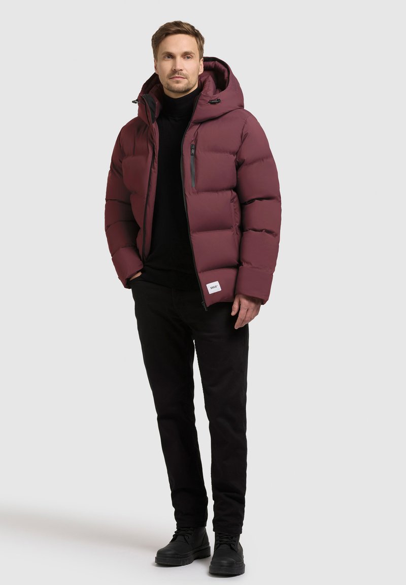 Bordeaux pufferjakke med hætte, lynlås foran og sidelommer. Båret over en sort turtleneck og sorte bukser, med sorte sko.