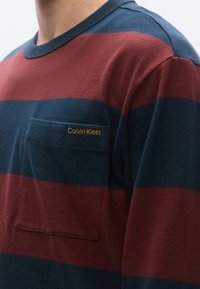 Ριγέ πουκάμισο με μακριά μανίκια σε σκούρο μπλε και μαρουνοκόκκινο χρώμα, με στρογγυλή λαιμόκοψη και μπροστινή τσέπη στο στήθος με κεντημένο το "Calvin Klein" σε χρυσό.