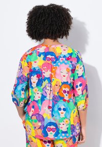 Blusa colorata con un motivo di volti stilizzati con vari stili di capelli e occhiali da sole. I materiali sembrano leggeri con maniche a 3/4.