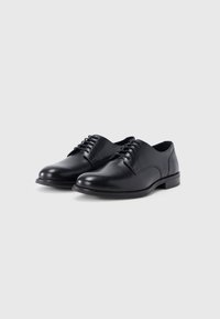 Paire de chaussures habillées pour homme en cuir noir avec lacets sur un fond gris clair uni.