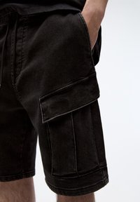Shorts cargo en denim noir avec une poche latérale, une coupe décontractée et des ourlets retroussés. Le tissu présente une légère texture avec des coutures visibles.