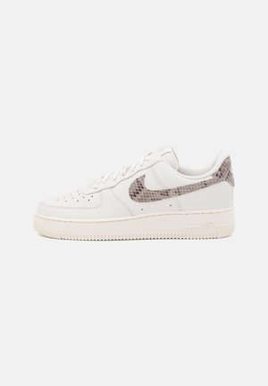 Białe niskie sneakersy z beżową podeszwą i wzorem wężowej skóry na logo Nike oraz akcentem na pięcie, widok z boku na jednolitym tle.