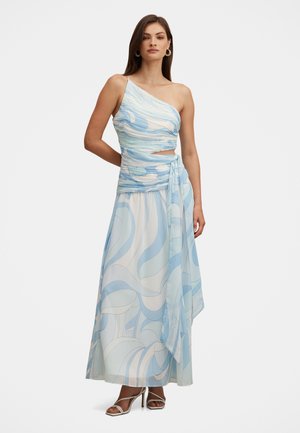 Donna che indossa un abito lungo monospalla con motivo blu e bianco, con una fascia legata in vita e sandali bianchi con tacco.