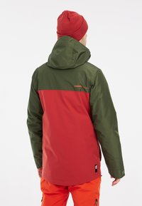 Rood en groene bicolor jas met een capuchon; gestructureerde stof; met een logo op de rug en een iets verlengde zoom; gecombineerd met oranje broek.