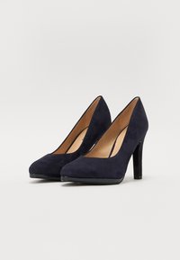 Marineblå semsket høyhælte pumps med spiss tå og en glatt, blank plattform. Hælhøyden er omtrent 10 cm.