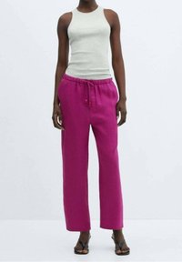 Lichtgrijze mouwloze top gecombineerd met levendige roze wijde broek met een trekkoord in de taille en subtiele textuur. Zwarte sandalen.