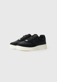Black low-top sneakers with perforated toe, black laces, red branding on tongue, and thick white rubber soles against gray background.